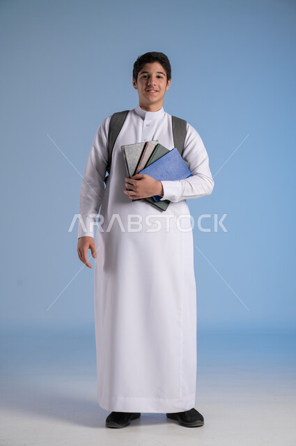 مفهوم الاجتهاد للوصول إلى النجاح، المناهج العلمية التعليمية، مفهوم العودة إلى المدارس، بورتريه لطالب عربي خليجي سعودي يرتدي الثوب التقليدي يحمل حقيبة الظهر المدرسية ويحمل مجموعه من الكتب، صورة لكامل الجسم، خلفية زرقاء