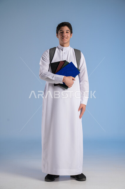 النظر الى الكاميرا بايماءات السرور، مفهوم الاجتهاد للوصول إلى النجاح، المناهج العلمية التعليمية، مفهوم العودة إلى المدارس، بورتريه لطالب عربي خليجي سعودي يرتدي الثوب التقليدي يحمل حقيبة الظهر المدرسية ويحمل مجموعه من الكتب، صورة لكامل الجسم، خلفية زرقاء