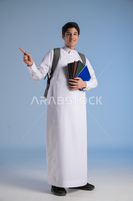 الاشارة باصبع السبابة الى شيء ما، مفهوم الاجتهاد للوصول إلى النجاح، المناهج العلمية التعليمية، مفهوم العودة إلى المدارس، بورتريه لطالب عربي خليجي سعودي يرتدي الثوب التقليدي يحمل حقيبة الظهر المدرسية ويحمل بيده كتاب، صورة لكامل الجسم، خلفية زرقاء