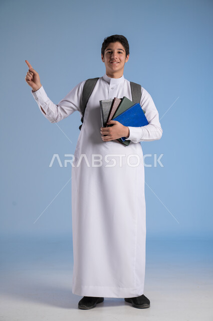 الاشارة باصبع السبابة الى شيء ما، مفهوم الاجتهاد للوصول إلى النجاح، المناهج العلمية التعليمية، مفهوم العودة إلى المدارس، بورتريه لطالب عربي خليجي سعودي يرتدي الثوب التقليدي يحمل حقيبة الظهر المدرسية ويحمل بيده كتاب، صورة لكامل الجسم، خلفية زرقاء