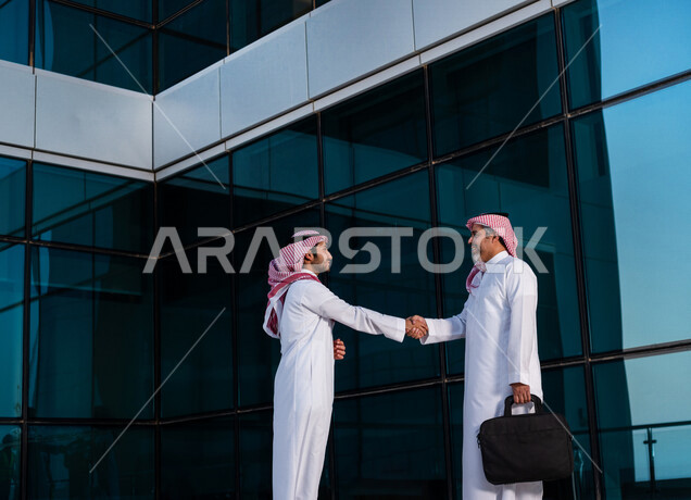 رجلان أعمال عربيان خليجيان سعوديان يرتديان الزي السعودي التقليدي يتصافحان داخل شركة سعودية ، اتفاق عمل ، عقد عمل جديد ، شراكة عمل
