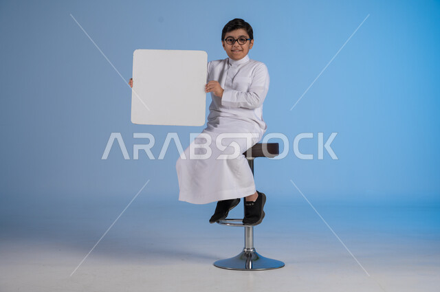 إستخدام لوحات توضيحية ووسائل تعليمية، إعلانات وعروضات تسويقية، بورتريه لشاب عربي سعودي خليجي يرتدي الثوب التقليدي يجلس على كرسي يحمل بيده لوحه بيضاء مربعة، موك اب مربع أبيض، لوح إعلاني فارغ، صورة الجسم بالطول الكامل, خلفية زرقاء
