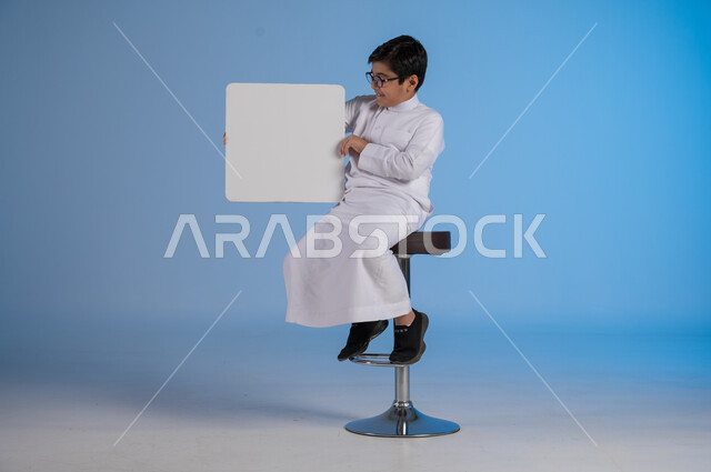 إستخدام لوحات توضيحية ووسائل تعليمية، إعلانات وعروضات تسويقية، بورتريه لشاب عربي سعودي خليجي يرتدي الثوب التقليدي يجلس على كرسي يحمل بيده لوحه بيضاء مربعة، موك اب مربع أبيض، لوح إعلاني فارغ، صورة الجسم بالطول الكامل, خلفية زرقاء