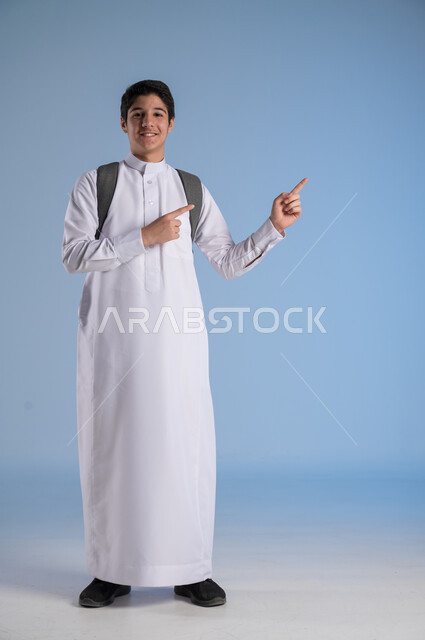 الاشارة الى شيء ما بايماءات السرور، متابعة الدروس والواجبات، تطوير المناهج التعليمية في السعودية, بورتريه لشاب عربي سعودي خليجي يرتدي الثوب التقليدي يحمل الحقيبة المدرسية, بداية العام الدراسي الجديد, أهمية التعليم في بناء المستقبل, صورة الجسم بالطول الكامل، خلفية زرقاء