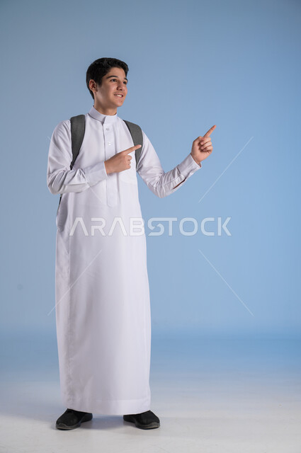 الاشارة الى شيء ما بايماءات السرور، متابعة الدروس والواجبات، تطوير المناهج التعليمية في السعودية, بورتريه لشاب عربي سعودي خليجي يرتدي الثوب التقليدي يحمل الحقيبة المدرسية, بداية العام الدراسي الجديد, أهمية التعليم في بناء المستقبل, صورة الجسم بالطول الكامل، خلفية زرقاء