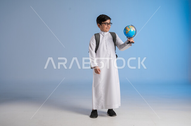مفهوم البحث والاستكشاف حول العالم, بورتريه لطفل عربي خليجي سعودي يرتدي الثوب التقليدي والنظارات الطبية يحمل مجسم الكرة الارضية, الاشارة إلى المجسم والنظر إلى الكاميرا, خريطة العالم الكروية, صورة الجسم بالطول الكامل, خلفية زرقاء
