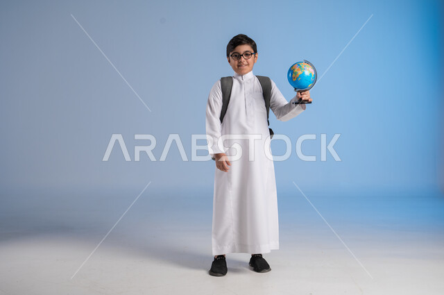 مفهوم البحث والاستكشاف حول العالم, بورتريه لطفل عربي خليجي سعودي يرتدي الثوب التقليدي والنظارات الطبية يحمل مجسم الكرة الارضية, الاشارة إلى المجسم والنظر إلى الكاميرا, خريطة العالم الكروية, صورة الجسم بالطول الكامل, خلفية زرقاء