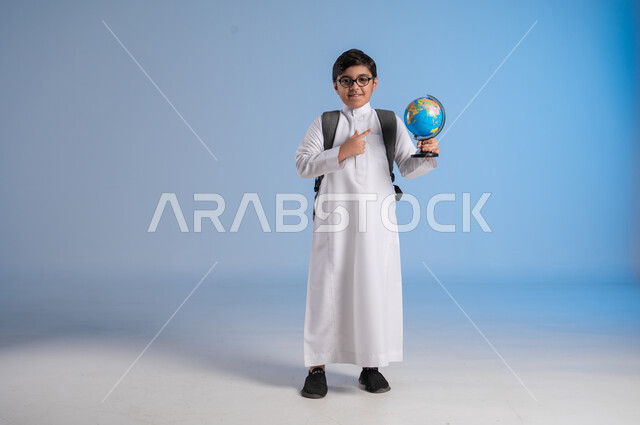 مفهوم البحث والاستكشاف حول العالم, بورتريه لطفل عربي خليجي سعودي يرتدي الثوب التقليدي والنظارات الطبية يحمل مجسم الكرة الارضية, الاشارة إلى المجسم والنظر إلى الكاميرا, خريطة العالم الكروية, صورة الجسم بالطول الكامل, خلفية زرقاء
