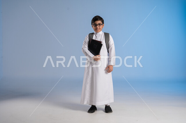 مفهوم الاجتهاد للوصول إلى النجاح، المناهج العلمية التعليمية، مفهوم العودة إلى المدارس، بورتريه لطالب عربي خليجي سعودي يرتدي الثوب التقليدي يحمل حقيبة الظهر المدرسية ويحمل بيده كتاب، صورة لكامل الجسم، خلفية زرقاء
