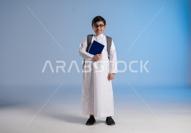 مراجعة الدروس وحل الواجبات، مفهوم الاجتهاد للوصول إلى النجاح، المناهج العلمية التعليمية، مفهوم العودة إلى المدارس، بورتريه لطالب عربي خليجي سعودي يرتدي الثوب التقليدي يحمل حقيبة الظهر المدرسية ويحمل بيده كتاب، صورة لكامل الجسم، خلفية زرقاء