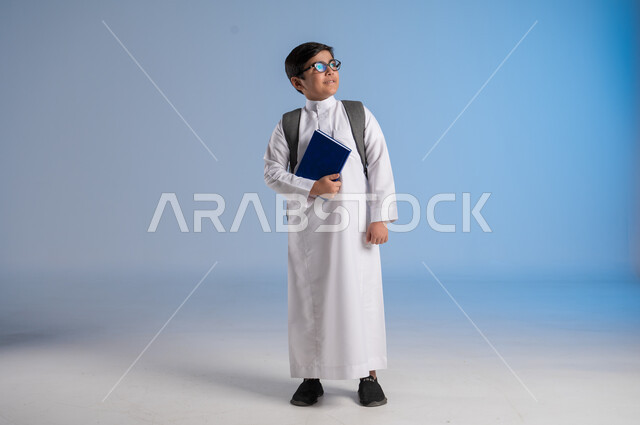 مفهوم الاجتهاد للوصول إلى النجاح، المناهج العلمية التعليمية، مفهوم العودة إلى المدارس، بورتريه لطالب عربي خليجي سعودي يرتدي الثوب التقليدي يحمل حقيبة الظهر المدرسية ويحمل بيده كتاب، صورة لكامل الجسم، خلفية زرقاء