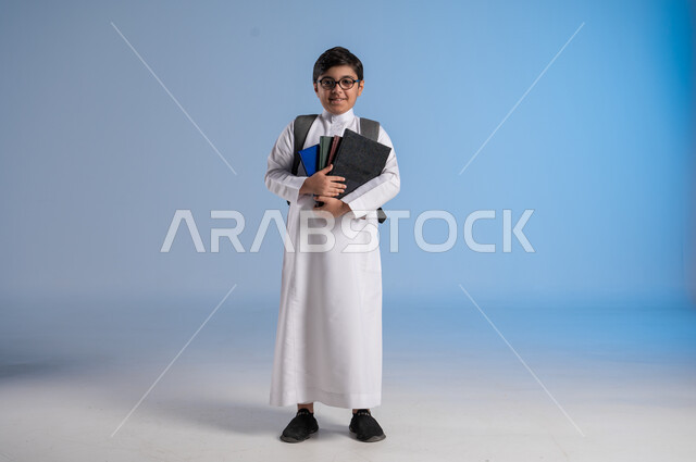 مفهوم الاجتهاد للوصول إلى النجاح، المناهج العلمية التعليمية، مفهوم العودة إلى المدارس، بورتريه لطالب عربي خليجي سعودي يرتدي الثوب التقليدي يحمل حقيبة الظهر المدرسية ويحمل بيده كتاب، صورة لكامل الجسم، خلفية زرقاء