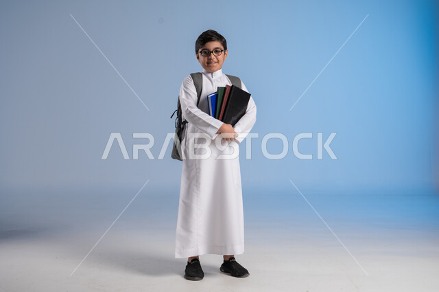 Reviewing lessons and solving homework, the concept of hard work to achieve success, educational scientific curricula, the concept of returning to school, a portrait of a Saudi Arabian Gulf student wearing traditional dress carrying a school backpack and holding a book in his hand, a full body image, blue background