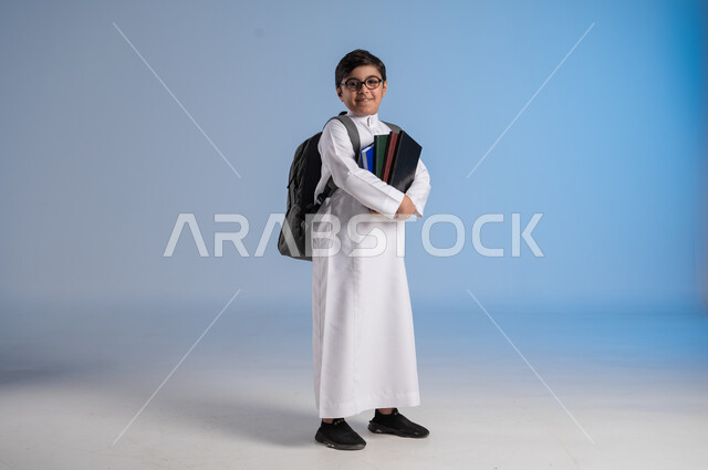 مفهوم الاجتهاد للوصول إلى النجاح، المناهج العلمية التعليمية، مفهوم العودة إلى المدارس، بورتريه لطالب عربي خليجي سعودي يرتدي الثوب التقليدي يحمل حقيبة الظهر المدرسية ويحمل بيده كتاب، صورة لكامل الجسم، خلفية زرقاء