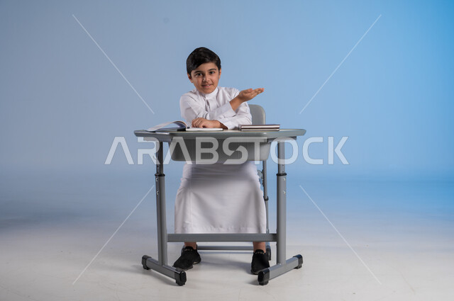 Pointing with his hand to something, seriousness and diligence in studying and reviewing in order to excel, portrait of a Saudi Arabian Gulf child wearing the traditional white dress sitting in his seat with a group of books in front of him, the importance of knowledge and learning, full-length body image, blue background