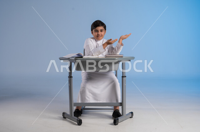 الاشارة بيده الى شيء ما، الجد والإجتهاد في الدراسة والمذاكرة من أجل التفوق, بورتريه لطفل عربي خليجي سعودي يرتدي الثوب الأبيض التقليدي يجلس في مقعده ويأمامه مجموعة من الكتب, أهمية العلم والتعلم,صورة الجسم بالطول الكامل، خلفية زرقاء