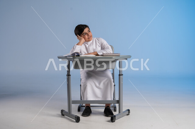التأمل والتفكير بشأن شيء ما، الجد والإجتهاد في الدراسة والمذاكرة من أجل التفوق, بورتريه لطفل عربي خليجي سعودي يرتدي الثوب الأبيض التقليدي يجلس في مقعده ويأمامه مجموعة من الكتب, أهمية العلم والتعلم,صورة الجسم بالطول الكامل، خلفية زرقاء