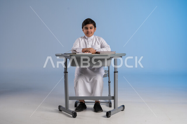 الجد والإجتهاد في الدراسة والمذاكرة من أجل التفوق, متابعة المناهج الدراسية بكل مثابرة وعزيمة, بورتريه لفتى عربي خليجي سعودي يرتدي الثوب التقليدي التقليدي يجلس في مقعده وأمامه مجموعة من الكتب, أهمية العلم والتعلم، خلفية زرقاء