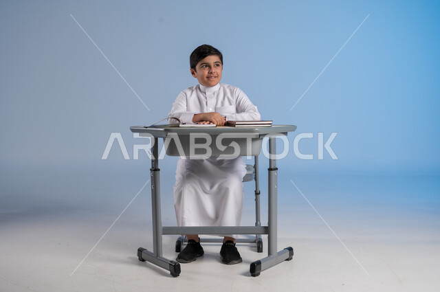 الجد والإجتهاد في الدراسة والمذاكرة من أجل التفوق, متابعة المناهج الدراسية بكل مثابرة وعزيمة, بورتريه لفتى عربي خليجي سعودي يرتدي الثوب التقليدي التقليدي يجلس في مقعده وأمامه مجموعة من الكتب, أهمية العلم والتعلم، خلفية زرقاء