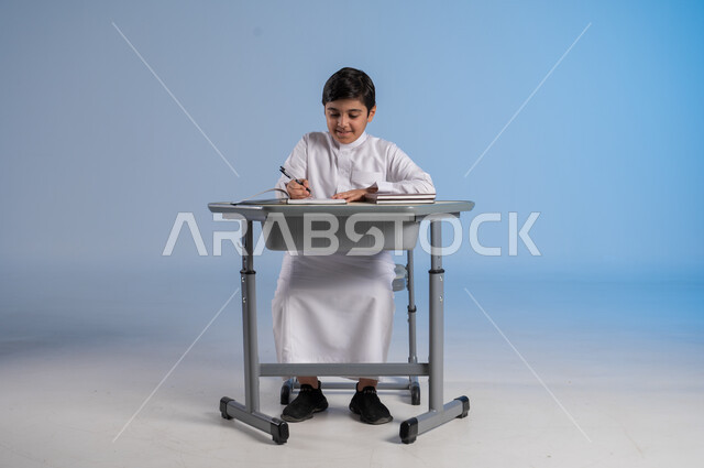 الجد والإجتهاد في الدراسة والمذاكرة من أجل التفوق, متابعة المناهج الدراسية بكل مثابرة وعزيمة, بورتريه لفتى عربي خليجي سعودي يرتدي الثوب التقليدي التقليدي يجلس في مقعده وأمامه مجموعة من الكتب, أهمية العلم والتعلم، خلفية زرقاء