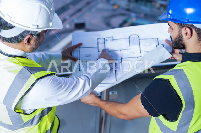 صورة مقربة من أعلى لمهندسان عربيان خليجيان سعوديان يرتديان خوذة و سترة خاصة بالعمل خلال مناقشة خطة تشييد موقع بناء و عمل أعلى سطح المبنى ، مع ورقة مخطط البناء ، اشراف و متابعة ، متابعة استراتيجية المشروع