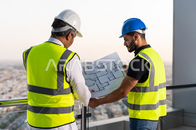 مهندسان عربيان خليجيان سعوديان يرتديان خوذة و سترة خاصة بالعمل خلال مناقشة خطة تشييد موقع بناء و عمل أعلى سطح المبنى ، مع ورقة مخطط البناء ، اشراف و متابعة ، متابعة استراتيجية المشروع