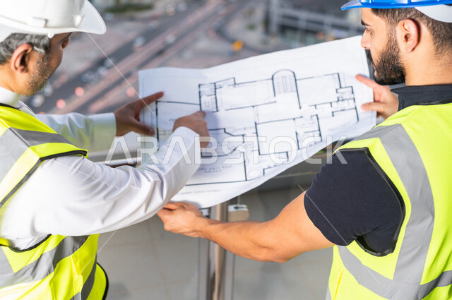 صورة مقربة من أعلى لمهندسان عربيان خليجيان سعوديان يرتديان خوذة و سترة خاصة بالعمل خلال مناقشة خطة تشييد موقع بناء و عمل أعلى سطح المبنى ، مع ورقة مخطط البناء ، اشراف و متابعة ، متابعة استراتيجية المشروع