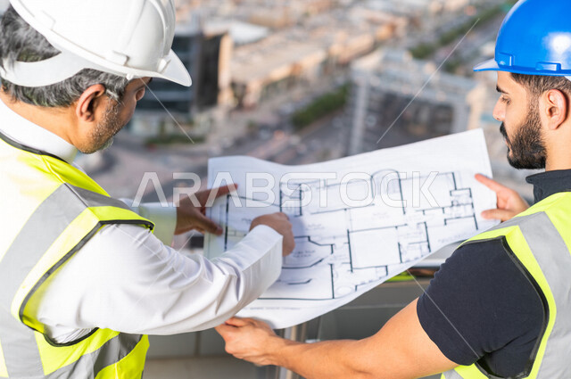 صورة مقربة من أعلى لمهندسان عربيان خليجيان سعوديان يرتديان خوذة و سترة خاصة بالعمل خلال مناقشة خطة تشييد موقع بناء و عمل أعلى سطح المبنى ، مع ورقة مخطط البناء ، اشراف و متابعة ، متابعة استراتيجية المشروع