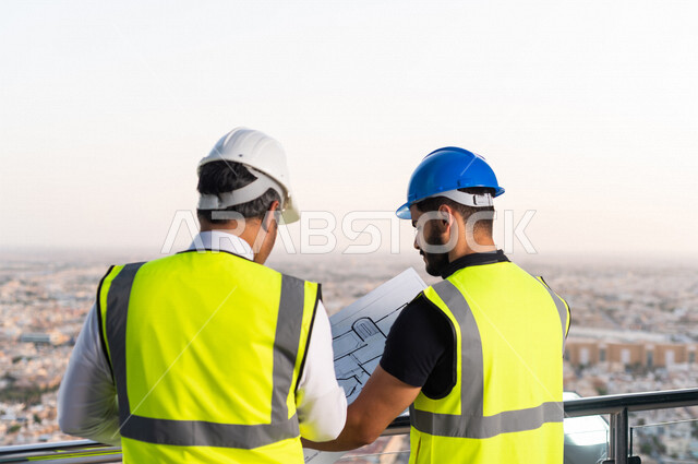 مهندسان عربيان خليجيان سعوديان يرتديان خوذة و سترة خاصة بالعمل خلال مناقشة خطة تشييد موقع بناء و عمل أعلى سطح المبنى ، مع ورقة مخطط البناء ، اشراف و متابعة ، متابعة استراتيجية المشروع