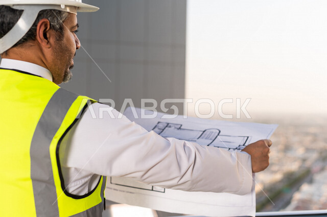 صورة مقربة لمهندس عربي خليجي سعودي يرتدي خوذة و سترة خاصة بالعمل أعلى سطح المبنى ، مع ورقة مخطط البناء ، اشراف و متابعة ، متابعة استراتيجية المشروع