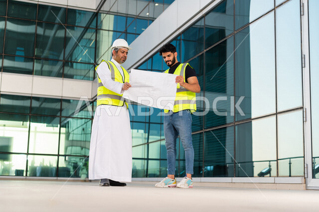 مهندسان عربيان خليجيان سعوديان يرتديان خوذة و سترة خاصة بالعمل خلال مناقشة خطة تشييد موقع بناء و عمل ، مع ورقة مخطط البناء ، اشراف و متابعة ، متابعة استراتيجية المشروع
