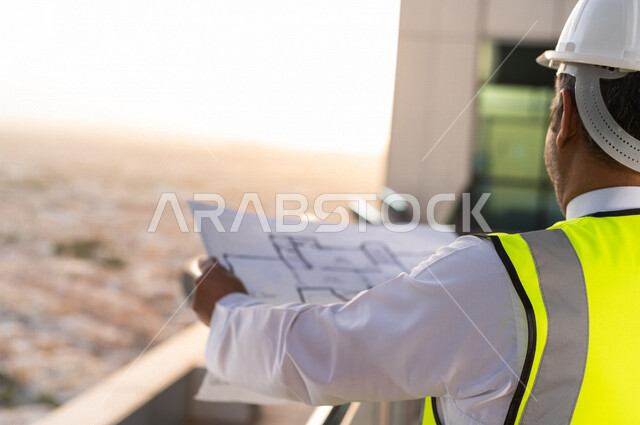 صورة مقربة لمهندس عربي خليجي سعودي يرتدي خوذة و سترة خاصة بالعمل أعلى سطح المبنى ، مع ورقة مخطط البناء ، اشراف و متابعة ، متابعة استراتيجية المشروع