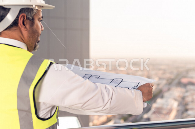صورة مقربة لمهندس عربي خليجي سعودي يرتدي خوذة و سترة خاصة بالعمل أعلى سطح المبنى ، مع ورقة مخطط البناء ، اشراف و متابعة ، متابعة استراتيجية المشروع