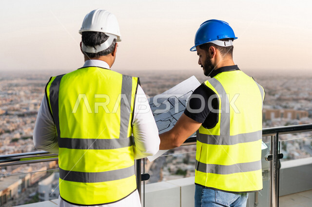 مهندسان عربيان خليجيان سعوديان يرتديان خوذة و سترة خاصة بالعمل خلال مناقشة خطة تشييد موقع بناء و عمل أعلى سطح المبنى ، مع ورقة مخطط البناء ، اشراف و متابعة ، متابعة استراتيجية المشروع