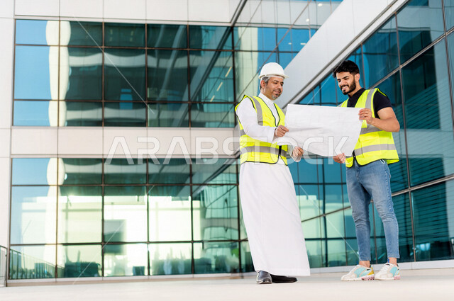مهندسان عربيان خليجيان سعوديان يرتديان خوذة و سترة خاصة بالعمل خلال مناقشة خطة تشييد موقع بناء و عمل ، مع ورقة مخطط البناء ، اشراف و متابعة ، متابعة استراتيجية المشروع