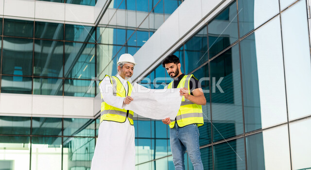 مهندسان عربيان خليجيان سعوديان يرتديان خوذة و سترة خاصة بالعمل خلال مناقشة خطة تشييد موقع بناء و عمل ، مع ورقة مخطط البناء ، اشراف و متابعة ، متابعة استراتيجية المشروع