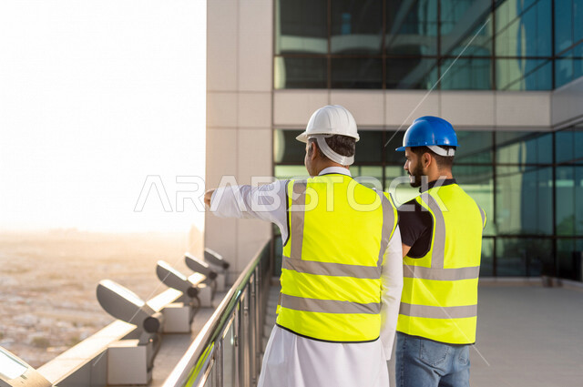 مهندسان عربيان خليجيان سعوديان يرتديان خوذة و سترة خاصة بالعمل خلال مناقشة خطة تشييد موقع بناء و عمل أعلى سطح المبنى ، مع ورقة مخطط البناء ، اشراف و متابعة ، متابعة استراتيجية المشروع