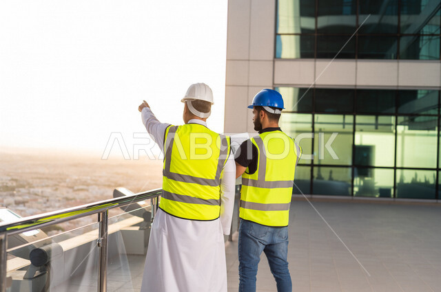 مهندسان عربيان خليجيان سعوديان يرتديان خوذة و سترة خاصة بالعمل خلال مناقشة خطة تشييد موقع بناء و عمل أعلى سطح المبنى ، مع ورقة مخطط البناء ، اشراف و متابعة ، متابعة استراتيجية المشروع