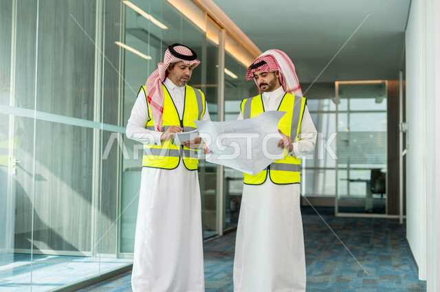 مهندسان معماريان سعوديان خليجيان يحملان مخطط لمشروع البناء للتخطيط وتنفيذ العمل في داخل المكتب في المملكة العربية السعودية, إرتداء سترة الحماية الخاصة بالعمل, تدقيق العمل، الإشراف والنقاش في العمل، مفهوم الهندسة، مهن ووظائف هندسية