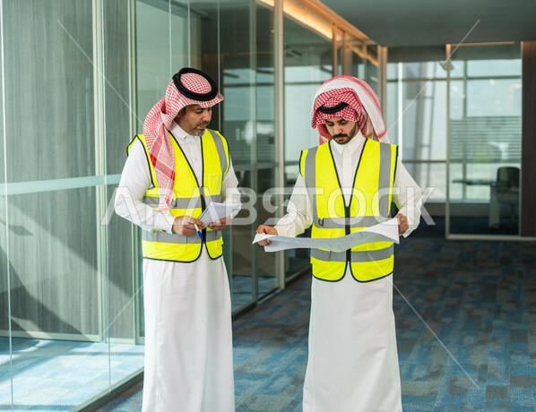 Two Saudi Gulf architects holding a blueprint for a construction project to plan and implement work inside the office in the Kingdom of Saudi Arabia, wearing protective work jackets, work auditing, supervision and discussion at work, engineering concept,