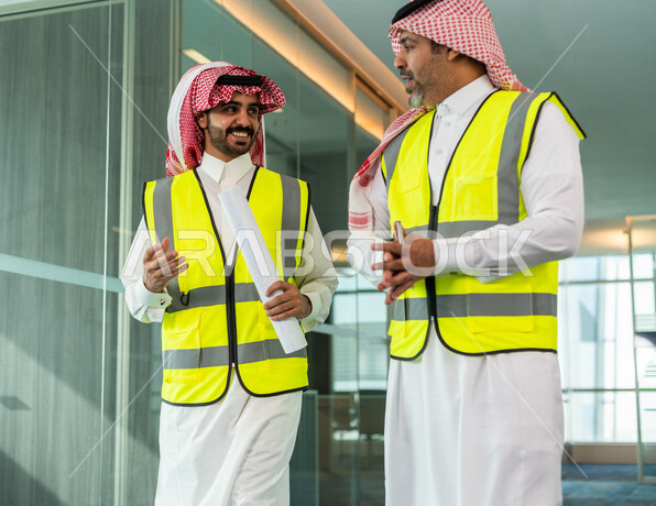 مهندسان معماريان سعوديان خليجيان يحملان مخطط لمشروع البناء للتخطيط وتنفيذ العمل في داخل المكتب في المملكة العربية السعودية, إرتداء سترة الحماية الخاصة بالعمل, تدقيق العمل، الإشراف والنقاش في العمل، مفهوم الهندسة، مهن ووظائف هندسية