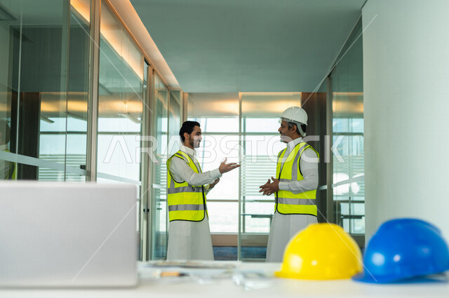 صورة مقربة لمكتب هندسي بخلفية مهندسان سعوديان خليجيان، يتناقشان لبدء مشروع جديد، استخدام جهاز الحاسوب المحمول، العمل في مجال الهندسة في داخل المكتب في المملكة العربية السعودية, إرتداء سترة الحماية الخاصة بالعمل، مهن ووظائف هندسية، اجتماع فريق عمل خبير