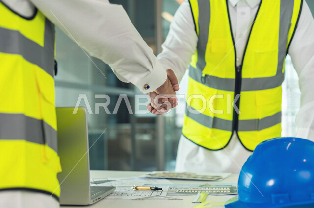 صورة مقربة لمهندسان معماريان سعوديان خليجيان ، يتصافحان لبدء مشروع جديد، العمل في مجال الهندسة في داخل المكتب في المملكة العربية السعودية, إرتداء سترة الحماية الخاصة بالعمل، الإشراف والنقاش، مهن ووظائف هندسية، فريق عمل خبير، خطط ومشاريع معمارية