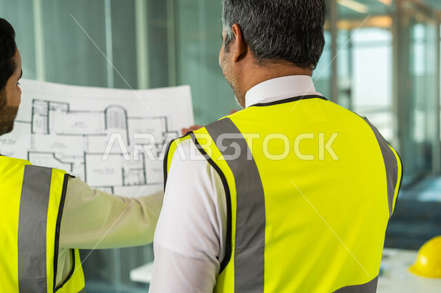 مهندسان معماريان سعوديان خليجيان يحملان مخطط لمشروع البناء للتخطيط وتنفيذ العمل في داخل المكتب في المملكة العربية السعودية, إرتداء سترة الحماية الخاصة بالعمل, تدقيق العمل، الإشراف والنقاش في العمل، مفهوم الهندسة، مهن ووظائف هندسية