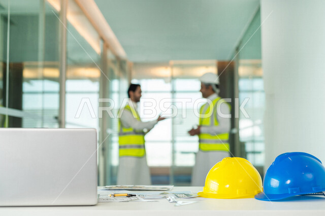 صورة مقربة لمكتب هندسي بخلفية مهندسان سعوديان خليجيان، يتناقشان لبدء مشروع جديد، استخدام جهاز الحاسوب المحمول، العمل في مجال الهندسة في داخل المكتب في المملكة العربية السعودية, إرتداء سترة الحماية الخاصة بالعمل، مهن ووظائف هندسية، اجتماع فريق عمل خبير