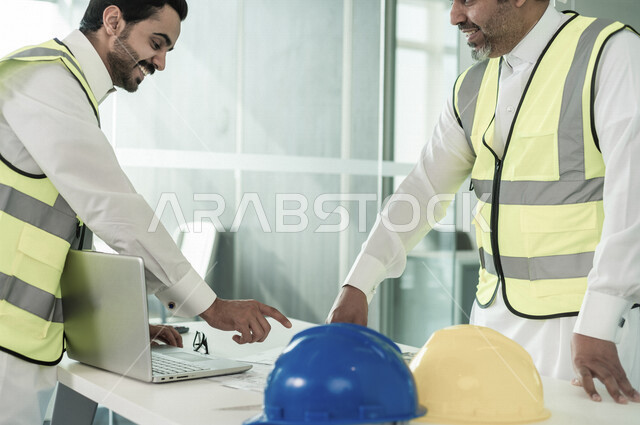 إجتماع مهندسان معماريان سعوديان خليجيان يعملان في مجال الهندسة المعماري في داخل المكتب في المملكة العربية السعودية, إرتداء سترة الحماية الخاصة بالعمل, تدقيق العمل، الإشراف والنقاش، مفهوم الهندسة، مهن ووظائف هندسية ، فريق عمل خبير، خطط ومشاريع معمارية