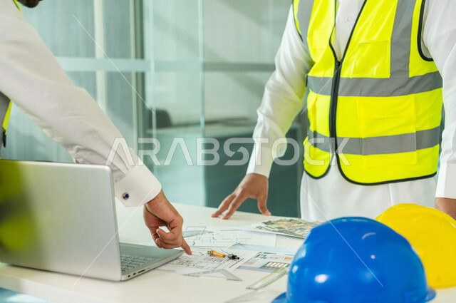 إجتماع مهندسان معماريان سعوديان خليجيان يعملان في مجال الهندسة المعماري في داخل المكتب في المملكة العربية السعودية, إرتداء سترة الحماية الخاصة بالعمل, تدقيق العمل، الإشراف والنقاش، مفهوم الهندسة، مهن ووظائف هندسية ، فريق عمل خبير، خطط ومشاريع معمارية