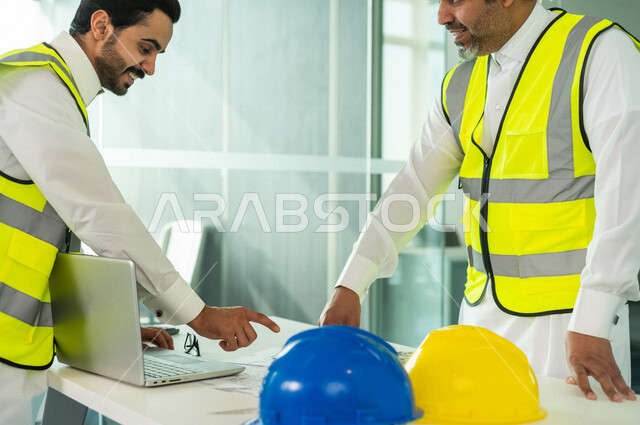 إجتماع مهندسان معماريان سعوديان خليجيان يعملان في مجال الهندسة المعماري في داخل المكتب في المملكة العربية السعودية, إرتداء سترة الحماية الخاصة بالعمل, تدقيق العمل، الإشراف والنقاش، مفهوم الهندسة، مهن ووظائف هندسية ، فريق عمل خبير، خطط ومشاريع معمارية