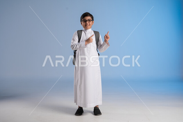 Academic schools in Saudi Arabia, preparing for the new school year, a Saudi Arabian Gulf student wearing traditional dress and glasses carrying a school backpack raising his hands up, gestures of enthusiasm and happiness for going back to school, full body image, blue background