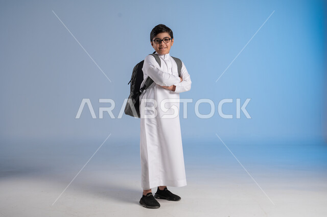 Academic schools in Saudi Arabia, preparing for the new school year, a Saudi Arabian Gulf student wearing traditional dress and glasses carrying a school backpack raising his hands up, gestures of enthusiasm and happiness for going back to school, full body image, blue background